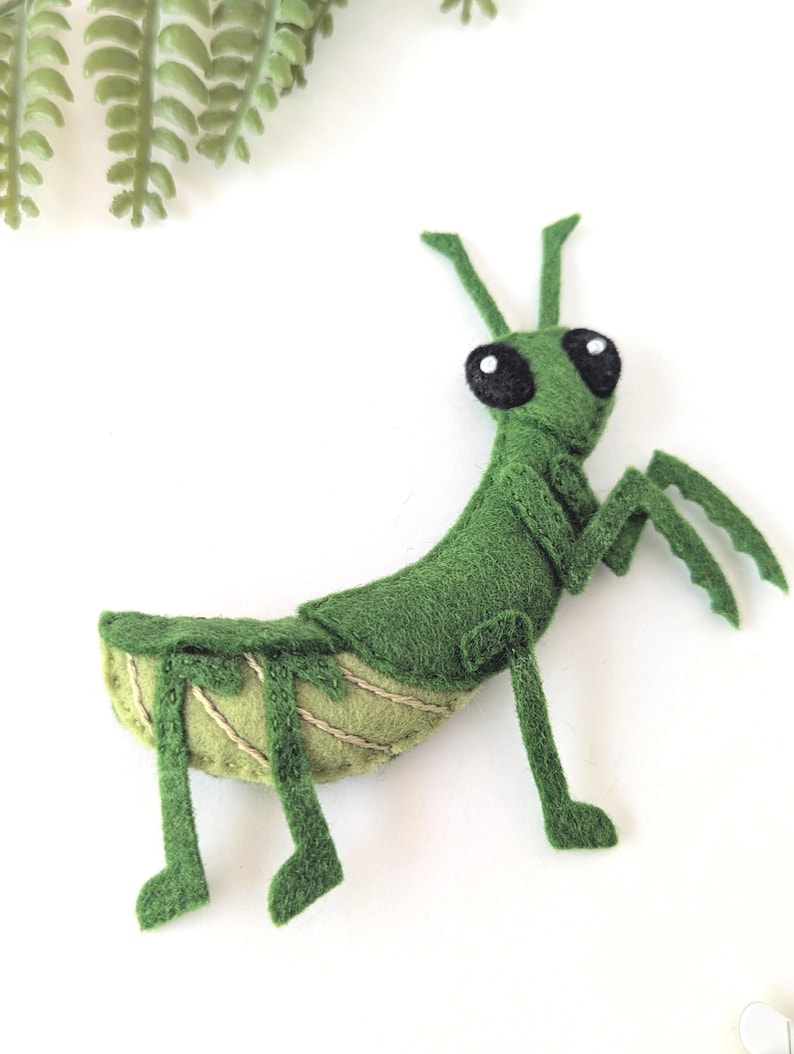 May include: A handmade green felt praying mantis toy with large black eyes and detailed legs. The insect is positioned on a white surface, with a sprig of green foliage in the upper left corner. The toy is a whimsical, handcrafted item.