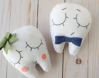 Puede incluir: Dos juguetes de peluche con forma de diente hechos de fieltro blanco. Un diente tiene un lazo verde y dos manchas de color melocotón. El otro diente tiene una pajarita azul.