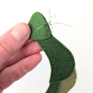May include: Close-up of a handmade felt leaf in various shades of green. The leaf is being sewn with a needle and thread. The felt has a soft texture, and the stitching is visible. The background is plain white.