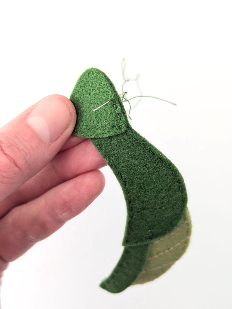 May include: Close-up of a handmade felt leaf in various shades of green. The leaf is being sewn with a needle and thread. The felt has a soft texture, and the stitching is visible. The background is plain white.