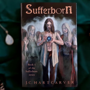May include: Book cover for "Sufferborn" by J.C. Hartcarver. The cover art features three figures with long hair and pale skin, with one holding a glowing orb. The title is in a copper-colored font, with the subtitle "Book 1 of the Sufferborn series".