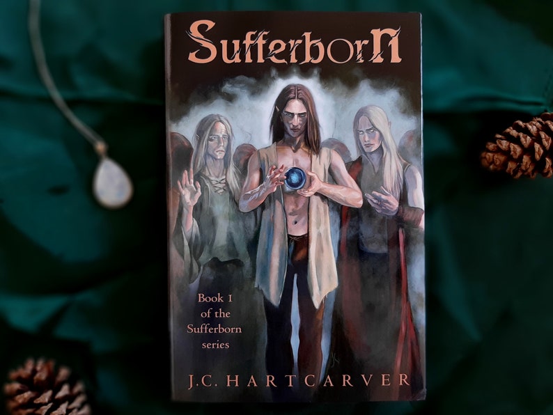 May include: Book cover for "Sufferborn" by J.C. Hartcarver. The cover art features three figures with long hair and pale skin, with one holding a glowing orb. The title is in a copper-colored font, with the subtitle "Book 1 of the Sufferborn series".