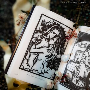 May include: An open book with black and white illustrations. The main illustration depicts two figures embracing. A silver necklace with a teardrop-shaped pendant rests on the page. The text "Kalea dreams sweetly of Dorhen" is visible.