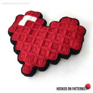 May include: A red crocheted heart-shaped pillow with a white square in the center. The pillow has a black crocheted border.