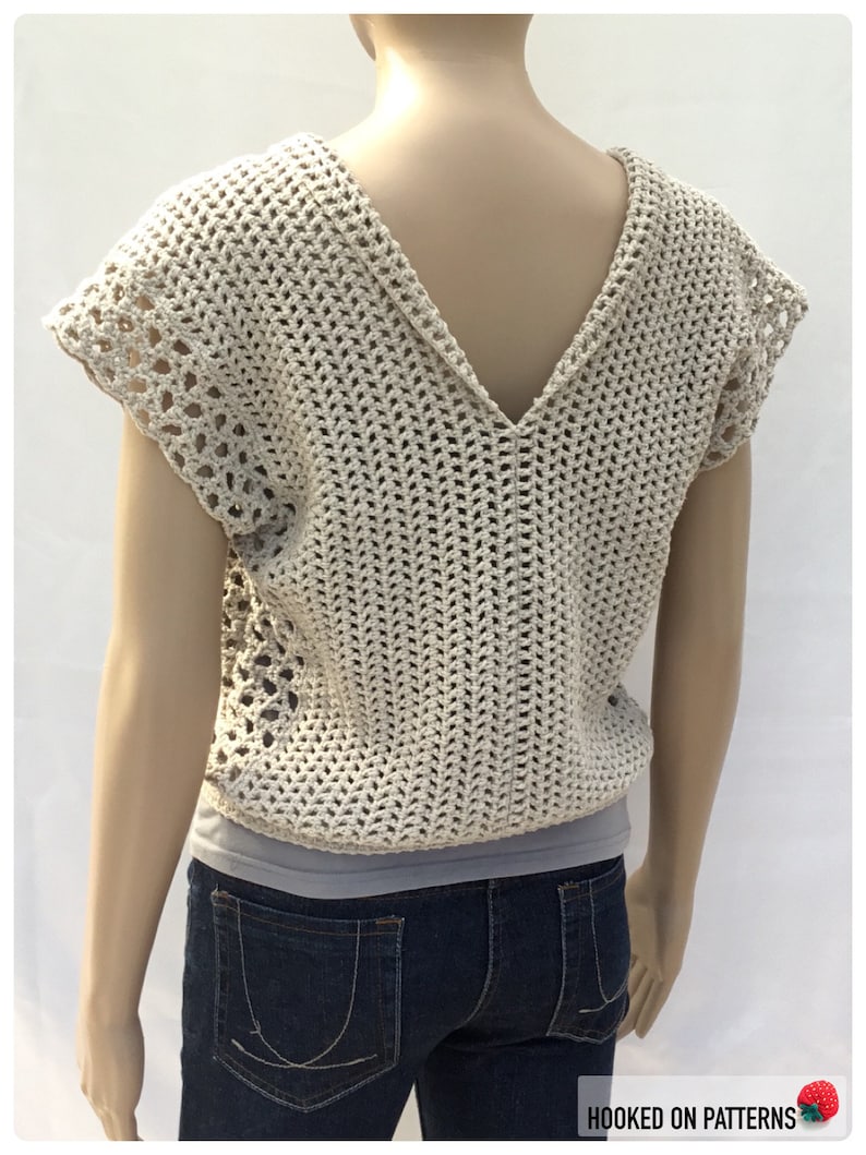 May include: A crocheted top with a V-neck and short sleeves. The top is made in a beige color and has a lacy pattern. The top is being worn by a mannequin.