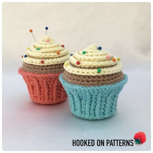 Op de afbeelding: Twee gehaakte cupcake-naaldenkussens, een met een lichtblauwe basis en de andere met een perzikbasis. Beide hebben een witte gehaakte frosting met kleurrijke sprinkles.