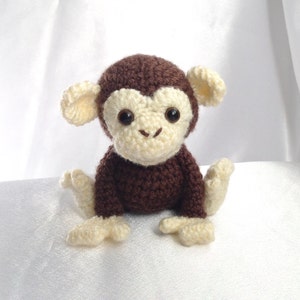 Puede incluir: Un juguete de mono de crochet marrón con cara y orejas de color crema. El mono está sentado sobre una superficie blanca.