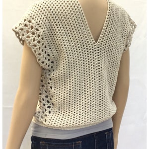 May include: A beige crochet top with a V-neck and short sleeves. The top has a lacy pattern and is worn over a white tank top and blue jeans.
