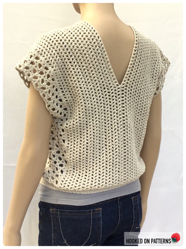 May include: A beige crochet top with a V-neck and short sleeves. The top has a lacy pattern and is worn over a white tank top and blue jeans.