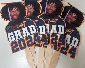 May include: Five graduation party decorations with a photo of a person wearing a graduation cap and gown. The decorations are orange, blue, and white with the text "GRAD 2024" on them.