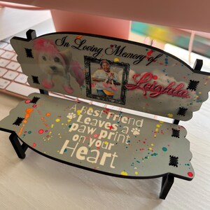 May include: A small wooden bench memorial with a gray background and colorful paint splatters. The bench has a photo of a dog and a person, and the text "In Loving Memory of Leighla A Best Friend Leaves a paw print on your Heart".