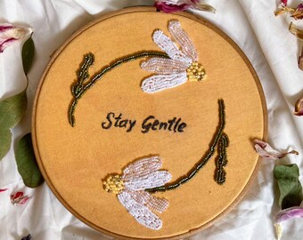 May include: A handmade embroidery hoop featuring two beaded daisy-like flowers and the phrase "Stay Gentle" in black thread on a mustard-colored fabric. The hoop is trimmed with a wooden frame and a green ribbon.