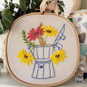 May include: Embroidered wall art featuring a blue coffee pot design with yellow and red flowers. The embroidery is set in a wooden hoop and includes a pink ribbon for hanging. The artwork is a charming piece of home decor.
