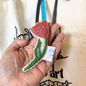 May include: A beaded tulip ornament with a red flower and green stem. The ornament is held in front of a cream-coloured tote bag with a black floral design and the word "art". A small tag is attached to the ornament.
