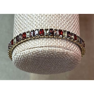 May include: A gold-toned bracelet featuring a continuous row of rectangular baguette-cut gemstones in alternating shades of deep red, clear white, and pale green, set in a metallic frame that curves along the display form.