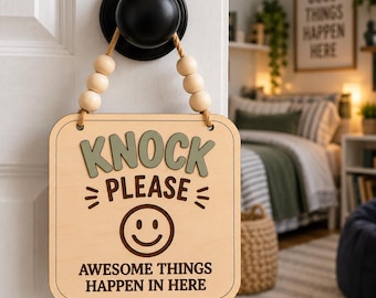 May include: A square wooden door sign hanging from a black knob by a jute rope with light-colored wooden beads. The sign features the text "KNOCK PLEASE" in green and brown, a brown smiley face icon, and the text "AWESOME THINGS HAPPEN IN HERE".