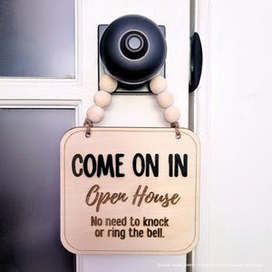 May include: A square light-colored wooden sign hanging from a black doorknob by a string of light wooden beads. The sign features the black text "COME ON IN", "Open House", and "No need to knock or ring the bell." printed on the front surface.