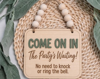 May include: A square wooden sign with rounded corners featuring the green text COME ON IN and brown text The Party's Waiting! No need to knock or ring the bell. The sign hangs from a jute rope adorned with eight light-colored wooden beads.