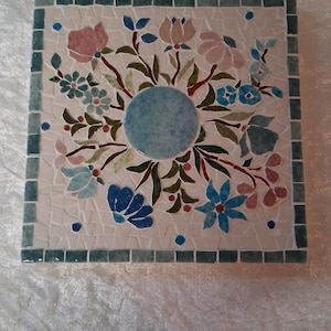 Könnte beinhalten: Quadratische Mosaikfliese mit floralem Design. Die Fliese zeigt einen zentralen hellblauen Kreis, umgeben von einem Mosaik aus weißen, blauen, rosa und grünen Fliesen. Der Rand ist ein dunkleres Türkis. Das Blumenmuster umfasst verschiedene Blumen und Blätter.