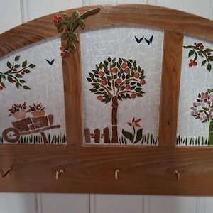 Puede incluir: Llavero de madera con tres paneles de mosaico que representan escenas de jardín. Los paneles presentan una carretilla con flores, un manzano y un árbol en flor con una luna creciente. El llavero tiene cuatro ganchos.