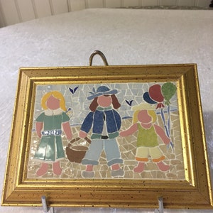 Puede incluir: Una obra de arte de mosaico enmarcada que representa a tres ni&ntilde;os, dos ni&ntilde;as y un ni&ntilde;o, caminando juntos. La ni&ntilde;a de la izquierda lleva un vestido verde, el ni&ntilde;o del medio lleva una chaqueta azul y pantalones vaqueros, y la ni&ntilde;a de la derecha lleva un vestido amarillo. El mosaico est&aacute; sobre un fondo azul y blanco con una cesta y globos.