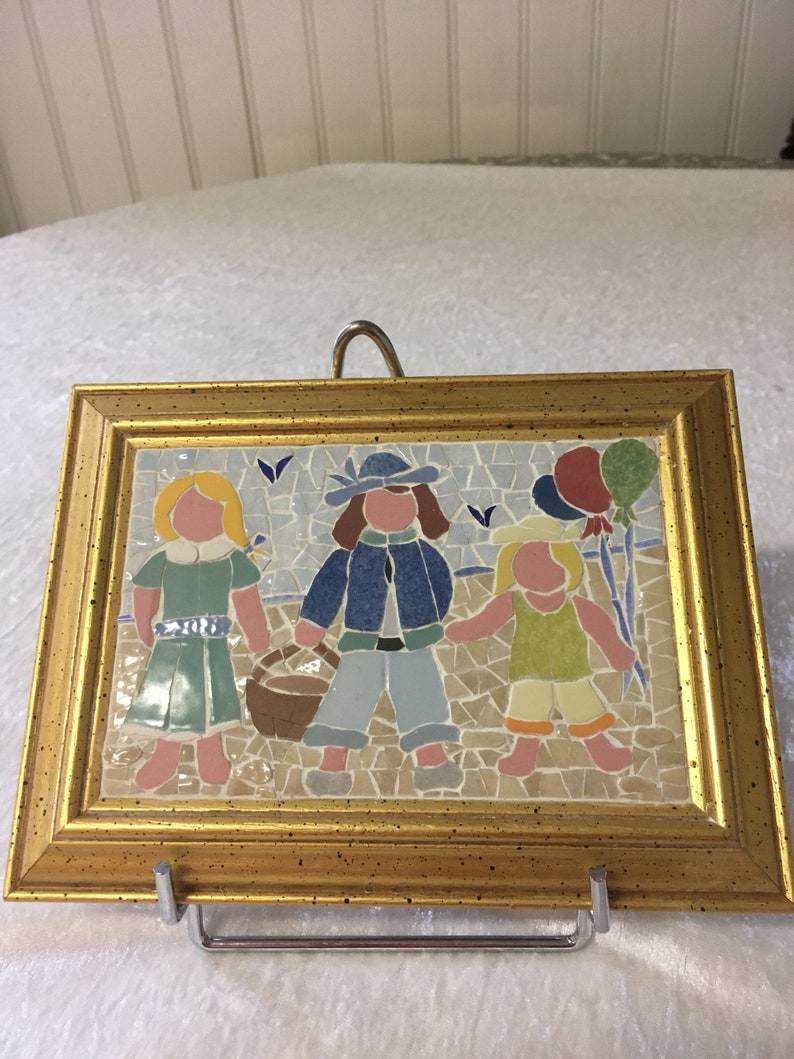 Puede incluir: Una obra de arte de mosaico enmarcada que representa a tres ni&ntilde;os, dos ni&ntilde;as y un ni&ntilde;o, caminando juntos. La ni&ntilde;a de la izquierda lleva un vestido verde, el ni&ntilde;o del medio lleva una chaqueta azul y pantalones vaqueros, y la ni&ntilde;a de la derecha lleva un vestido amarillo. El mosaico est&aacute; sobre un fondo azul y blanco con una cesta y globos.