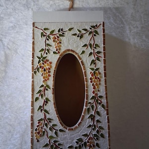 May include: A rectangular tissue box with a mosaic tile design. The box has an oval opening and is decorated with grape vines and clusters of grapes in green, brown, and yellow. A small string is attached at the top.