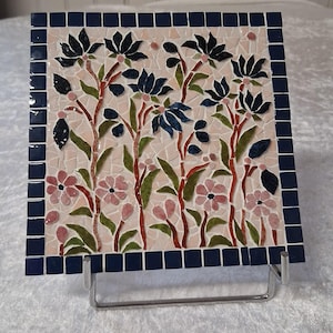 Könnte beinhalten: Quadratische Mosaikfliese mit floralem Design. Das Mosaik zeigt dunkelblaue Blumen, rosa Blumen und grüne Blätter auf hellrosa Hintergrund. Der Rand ist dunkelblau. Die Fliese wird auf einem Metallständer ausgestellt.