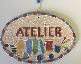 Placa de puerta de mosaico: "Atelier". Pequeñas herramientas de costura y botones sobre un soporte de madera pintada con cordón para colgar.