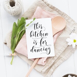 May include: A white card with black calligraphy that reads "This kitchen is for dancing". The card is on a folded linen napkin with a wooden spoon and a sprig of sage.