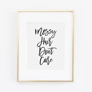 Puede incluir: Impresión enmarcada con el texto "Messy Hair Don't Care" en escritura negra. La impresión está sobre un fondo blanco y enmarcada en un marco dorado. La estética general es minimalista y moderna.
