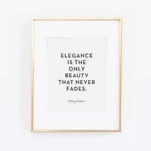May include: A white framed print with a gold frame. The print features a black and white quote that reads "Elegance is the only beauty that never fades." The quote is attributed to Audrey Hepburn.