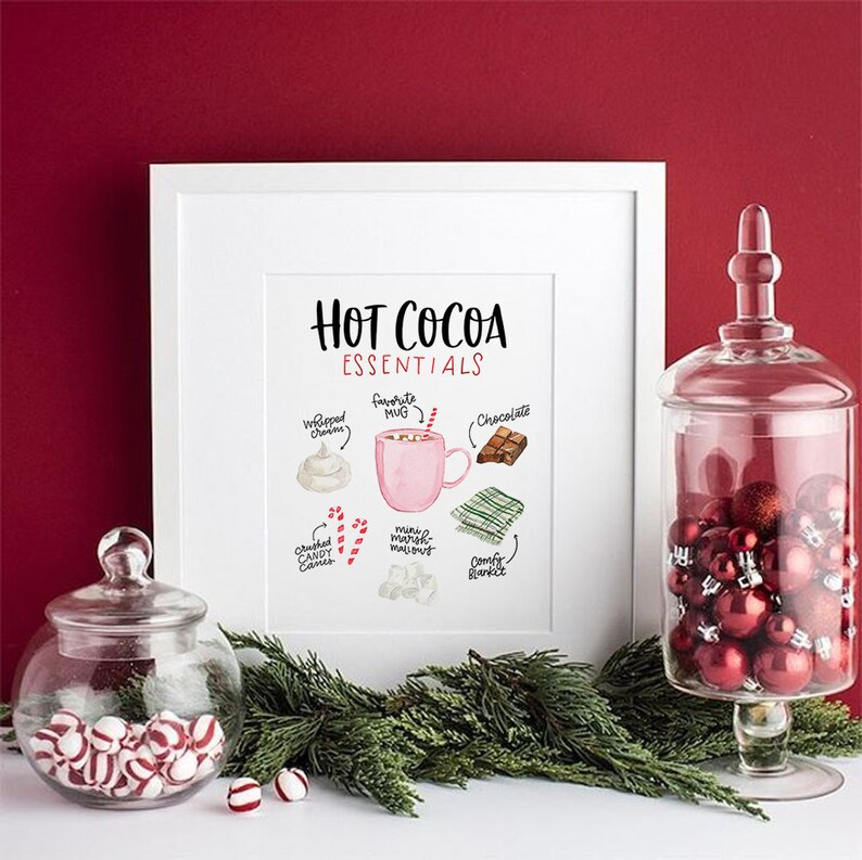 Puede incluir: Una impresi&oacute;n enmarcada en blanco con el texto "Hot Cocoa Essentials" e ilustraciones de ingredientes para chocolate caliente, incluyendo crema batida, una taza rosa, chocolate, mini malvaviscos, bastones de caramelo triturados y bastones de caramelo. La impresi&oacute;n est&aacute; sobre una mesa con un frasco de vidrio lleno de adornos rojos y un frasco de vidrio lleno de bastones de caramelo.