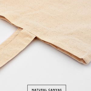 May include: Close-up of a natural canvas tote bag with a folded over edge. The bag is a light beige color and has a natural, unbleached look. The text "NATURAL CANVAS" is printed on the bag.