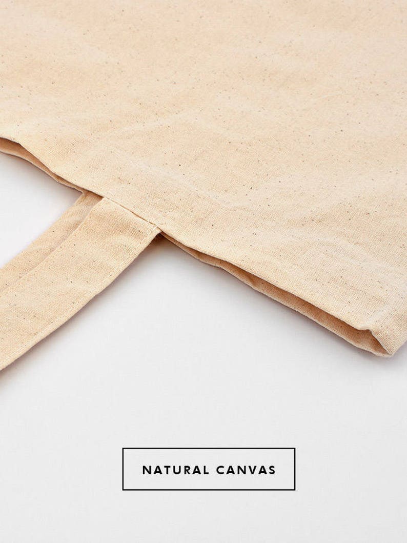May include: Close-up of a natural canvas tote bag with a folded over edge. The bag is a light beige color and has a natural, unbleached look. The text "NATURAL CANVAS" is printed on the bag.