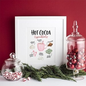 Puede incluir: Una impresi&oacute;n enmarcada en blanco con el texto "Hot Cocoa Essentials" e ilustraciones de ingredientes para chocolate caliente, incluyendo crema batida, una taza rosa, malvaviscos, chocolate, bastones de caramelo y una manta acogedora.