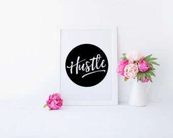 May include: A white framed print with a black circle and the word "Hustle" written in white. The print is sitting on a white surface with a vase of pink peonies.