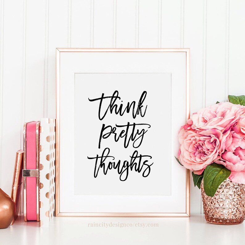 May include: A white framed print with the text "think pretty thoughts" in black script font. The print is on a white surface with a pink and gold polka dot notebook and a vase of pink flowers.