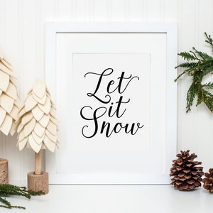 May include: White framed print with the text "Let it Snow" in black cursive script. The print is surrounded by two small white felt Christmas trees and a small green pine wreath. There are also three pine cones on a white surface.