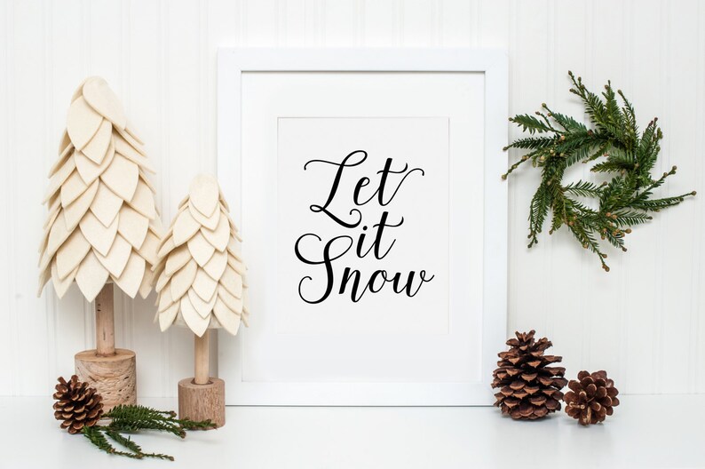 May include: White framed print with the text "Let it Snow" in black cursive script. The print is surrounded by two small white felt Christmas trees and a small green pine wreath. There are also three pine cones on a white surface.