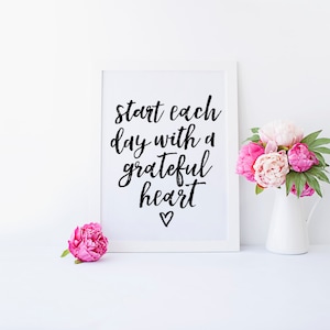 Puede incluir: Impresión enmarcada en blanco con el texto "Start each day with a grateful heart" y un pequeño símbolo de corazón. Flores rosas en una jarra blanca sobre una superficie blanca.