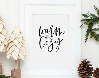 May include: White framed print with black calligraphy that reads "warm + cozy" on a white background. The print is sitting on a white surface with a small white pine tree on the left and a pine cone on the right.