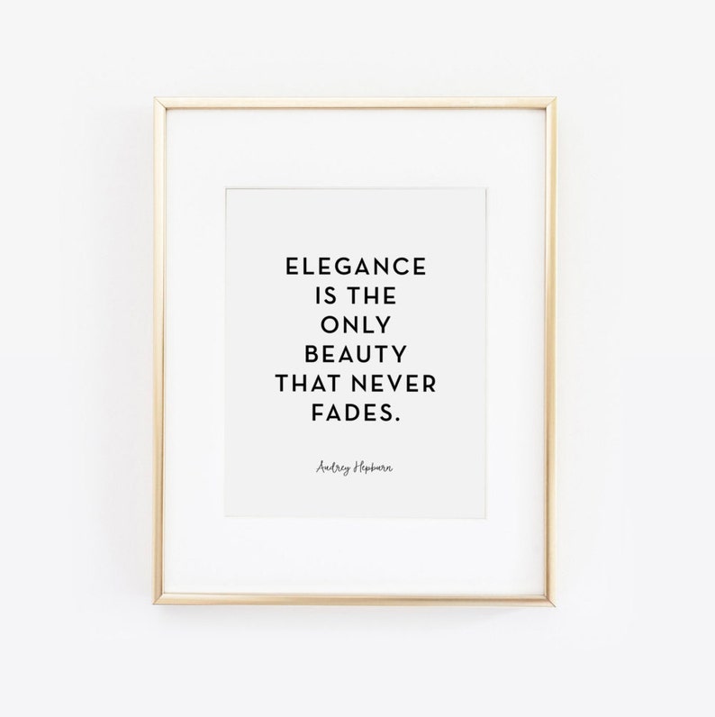 May include: A white framed print with a gold frame. The print features a black and white quote that reads "Elegance is the only beauty that never fades." The quote is attributed to Audrey Hepburn.