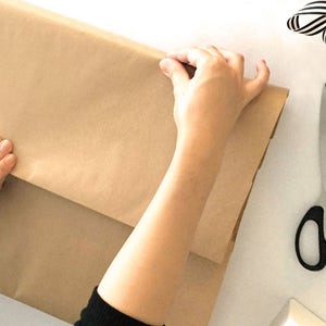 May include: A person wrapping a brown paper package with their hands. The paper has the text "rain city" printed on it. A pair of silver scissors with black handles are on the table.
