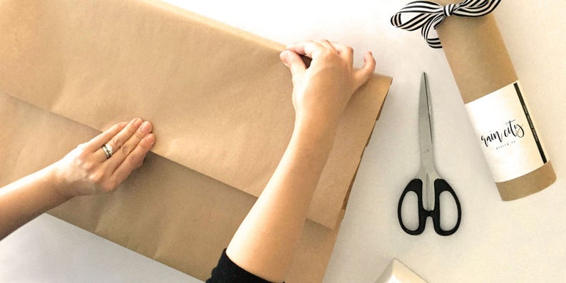 May include: A person wrapping a brown paper package with their hands. The paper has the text "rain city" printed on it. A pair of silver scissors with black handles are on the table.