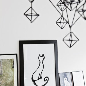 May include: A minimalist black and white cat art print in a black frame, displayed on a white shelf with other framed art. Above, a geometric black wire sculpture hangs against a white wall, creating a modern aesthetic.