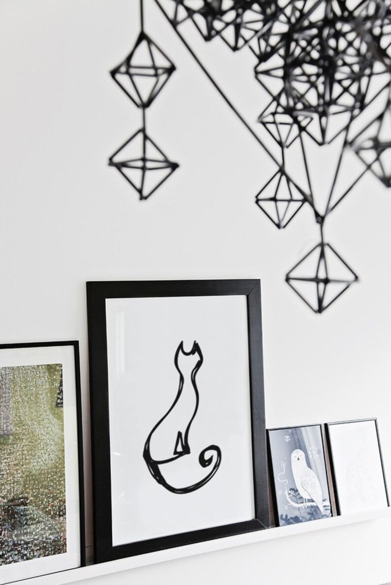 May include: A minimalist black and white cat art print in a black frame, displayed on a white shelf with other framed art. Above, a geometric black wire sculpture hangs against a white wall, creating a modern aesthetic.