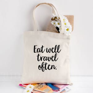 May include: A white canvas tote bag with black text that reads "Eat well travel often." The bag is sitting on a stack of magazines and a bouquet of white daisies.