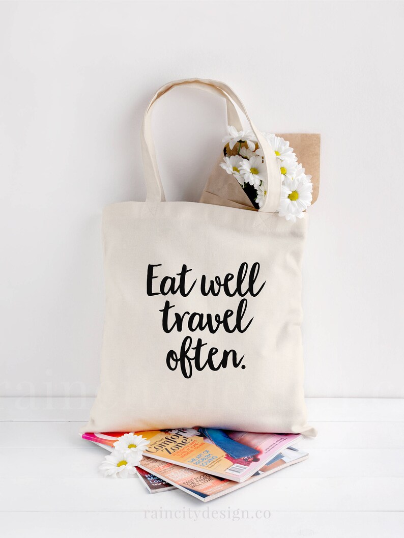 May include: A white canvas tote bag with black text that reads "Eat well travel often." The bag is sitting on a stack of magazines and a bouquet of white daisies.