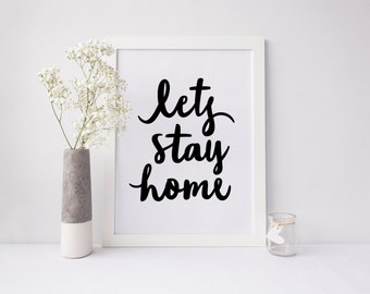 May include: White framed print with black calligraphy that reads "let's stay home". The print is on a white surface with a vase of white flowers and a clear glass jar.